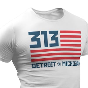 Puede incluir: Camiseta blanca con un diseño gráfico azul y rojo. El gráfico presenta el número "313" con un diseño de bandera estadounidense y el texto "DETROIT MICHIGAN".