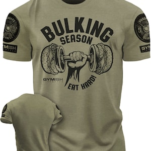 Peut inclure: Un t-shirt vert avec un motif noir d'une main soulevant une haltère avec deux pains à hamburger à chaque extrémité. Le texte "BULKING SEASON" est au-dessus de l'haltère et "EAT HARD!" est en dessous. Le texte "GYMISH LIFESTYLE" est sous l'haltère.