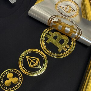 May include: A black t-shirt with gold foil iron-on designs of cryptocurrency logos. The logos include Bitcoin, Ethereum, and Ripple.