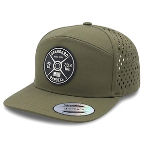 May include: An olive green snapback cap with a circular black patch on the front featuring the text STANDARD ATHL DEPT, 45 LB, 20.4 KG, and BARBELL with a small American flag. The side panels have laser-cut ventilation holes and a holographic sticker.