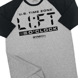 May include: A gray and black baseball tee shirt with the text "U.S. Time Zone LIFT O'CLOCK" and the logo "GYMISH LIFESTYLE".