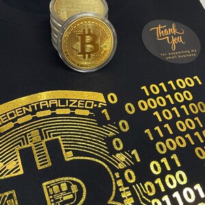 May include: Black t-shirt with a gold foil design of a circuit board and the text "DECENTRALIZED". The design also includes a gold foil Bitcoin symbol and binary code. The shirt has a thank you sticker with the text "Thank You for supporting my small business".
