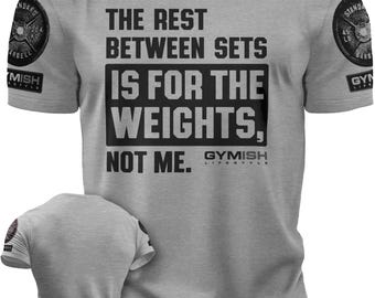 May include: Gray t-shirt with a black graphic that reads "The rest between sets is for the weights, not me." The graphic is surrounded by a black border. The shirt has a small logo on the left and right sleeves that reads "Gymish Lifestyle".