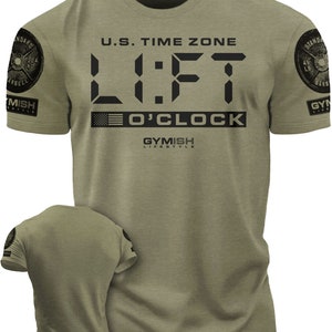 Może przedstawiać: Zielony T-shirt z cyfrowym wzorem zegara, który wyświetla "LIFT O'CLOCK" i tekst "U.S. TIME ZONE" nad zegarem. Koszulka ma małą grafikę amerykańskiej flagi pod zegarem i tekst "GYMISH LIFESTYLE" pod flagą. Koszulka ma na każdym rękawie grafikę czarno-białej płyty ciężarowej.