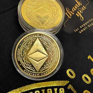 May include: Two gold-colored Ethereum Classic cryptocurrency coins in clear plastic cases. The coins feature a stylized Ethereum logo and the text "ethereum classic" and "open-source" and "decentralized".