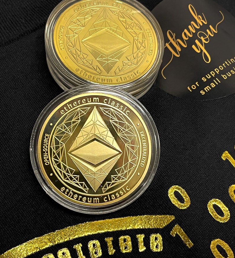 May include: Two gold-colored Ethereum Classic cryptocurrency coins in clear plastic cases. The coins feature a stylized Ethereum logo and the text "ethereum classic" and "open-source" and "decentralized".