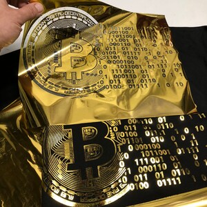 May include: A gold foil sheet with the text "FINE COPPER MAS MONETOF METALS BITCOIN DIGITAL DECENTRALIZED" and a stylized image of a Bitcoin logo. The foil sheet is being placed on top of a black fabric with a gold foil print of a Bitcoin logo.