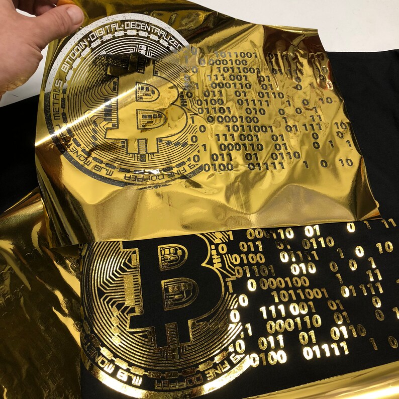 May include: A gold foil sheet with the text "FINE COPPER MAS MONETOF METALS BITCOIN DIGITAL DECENTRALIZED" and a stylized image of a Bitcoin logo. The foil sheet is being placed on top of a black fabric with a gold foil print of a Bitcoin logo.