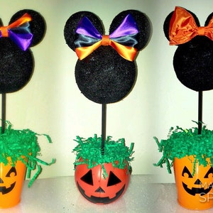 May include: Three Halloween-themed centerpieces featuring black Mickey Mouse ears with a bow on top. The ears are attached to a stick and placed in a small orange pumpkin-shaped container with green grass on top.