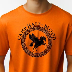 Op de afbeelding: Oranje T-shirt met een zwart Pegasus-embleem in een cirkelvormig ontwerp met de tekst "CAMP HALF-BLOOD". Het shirt met korte mouwen is gemaakt van een zachte stof.