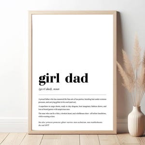 Op de afbeelding: Een ingelijste print met de woorden "girl dad" in vette zwarte letters. De definitie beschrijft een vaderfiguur die een prinsessenbeschermer, glitterkrijger en tutu-troubleshooter is. De print zit in een houten lijst.