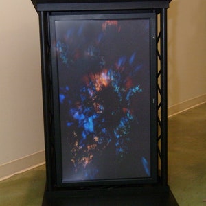 May include: A black stand with a black frame and a screen displaying a colorful abstract image. The screen is surrounded by a black frame and the stand has a black base.