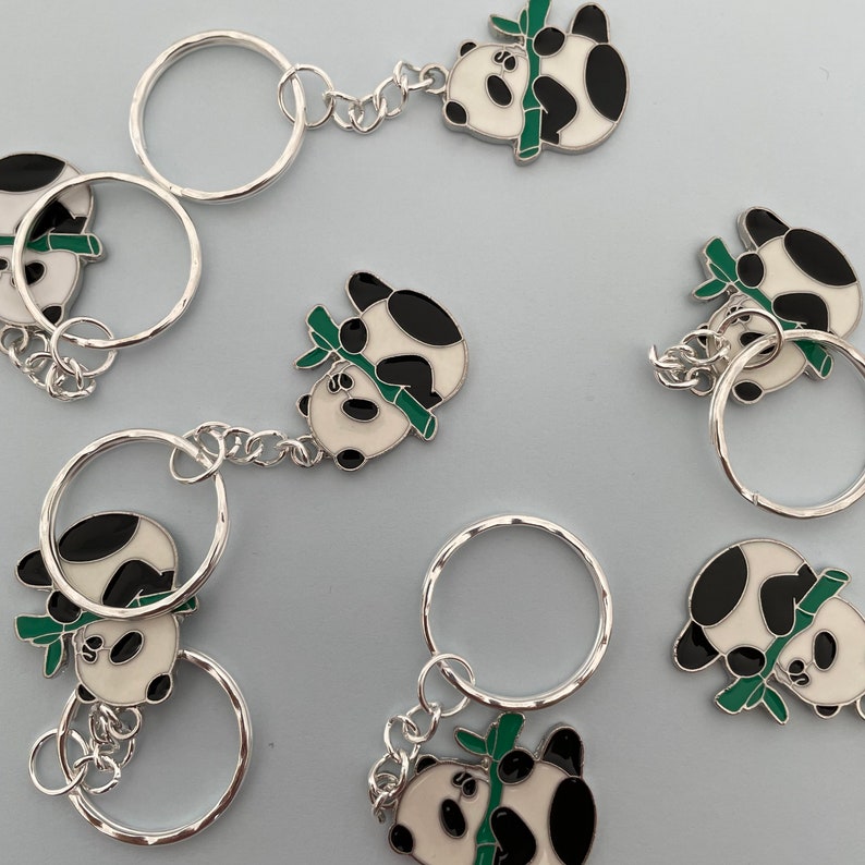 May include: A set of six keychains featuring panda bears holding bamboo stalks. The pandas are black and white with green bamboo stalks. The keychains are silver and have a circular shape.