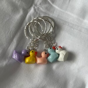 May include: Five colorful rubber duck keychains with silver key rings. The ducks are purple, yellow, pink, blue, and white.