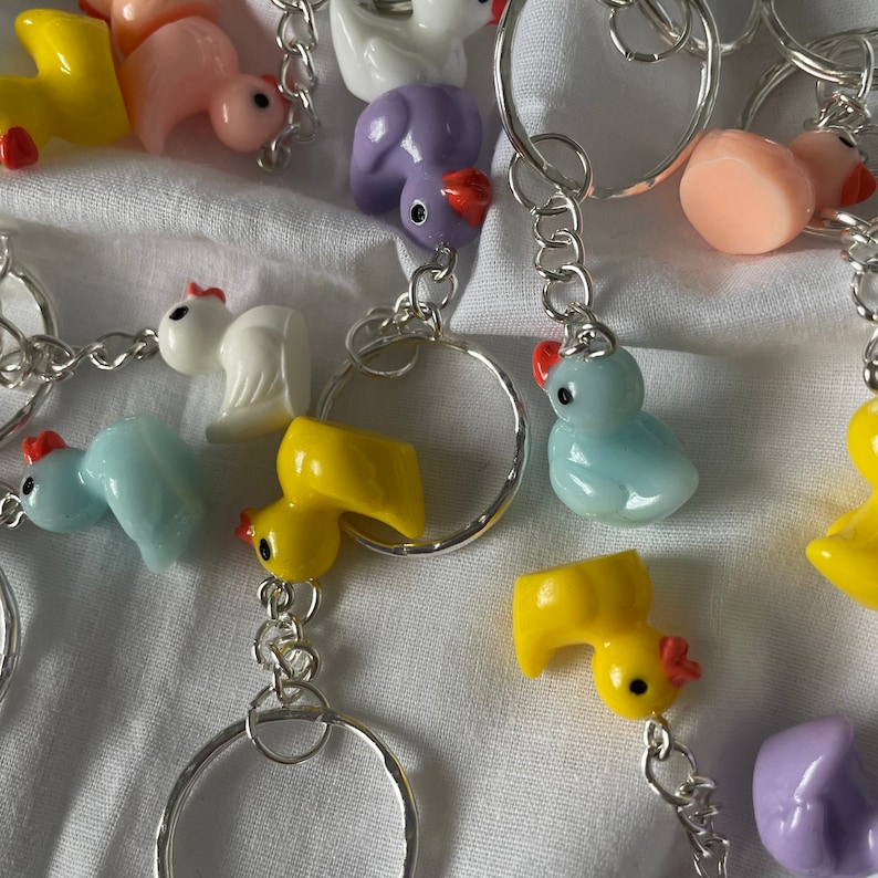 May include: A collection of colorful keychains featuring various animal designs, including ducks, chicks, and a purple bird. The keychains are attached to silver key rings and chains.