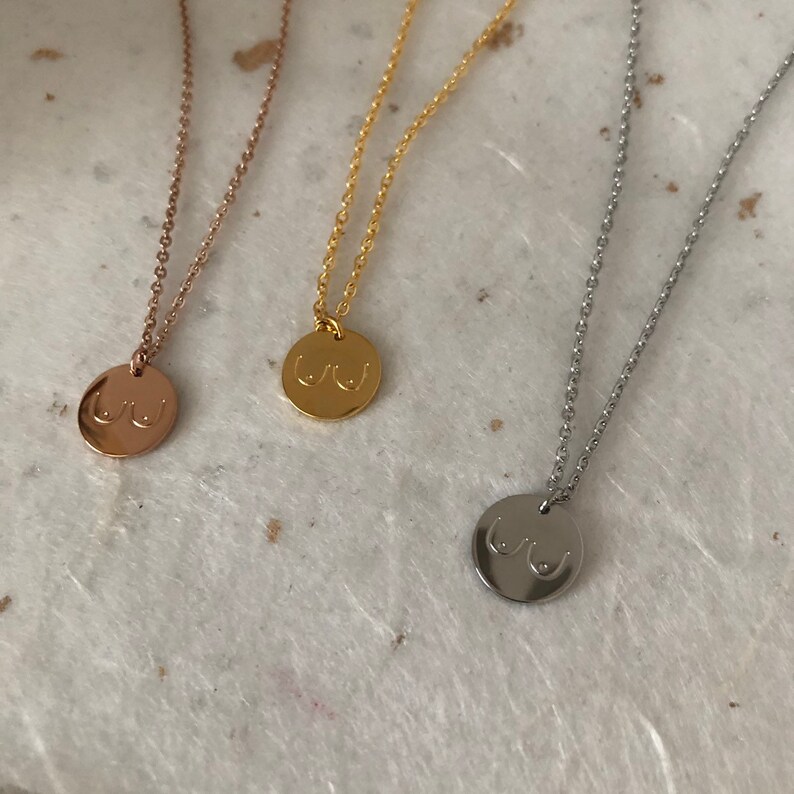 May include: Three necklaces with a round pendant featuring a stylized image of breasts. The necklaces are available in gold, rose gold, and silver.