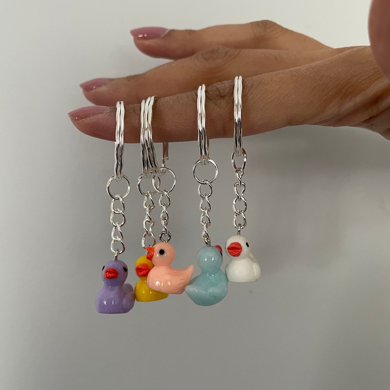 May include: A set of six colorful rubber duck keychains. The ducks are in shades of purple, yellow, pink, blue, and white. Each duck has a silver chain and ring attached.