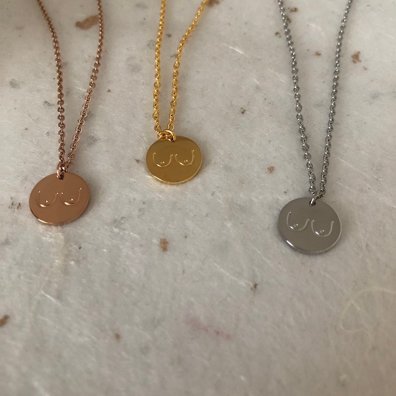 May include: Three necklaces with a round pendant featuring a stylized image of breasts. The necklaces are available in gold, rose gold, and silver.