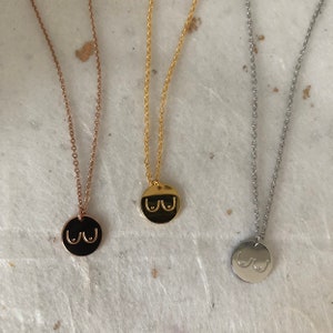 May include: Three necklaces with a round pendant featuring a stylized image of breasts. The necklaces are gold, rose gold, and silver.