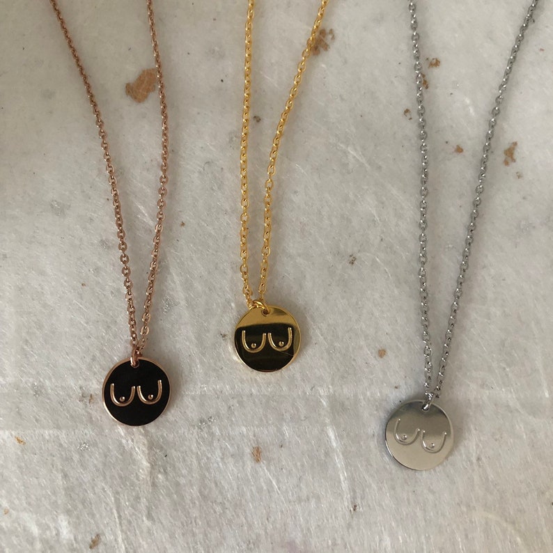 May include: Three necklaces with a round pendant featuring a stylized image of breasts. The necklaces are gold, rose gold, and silver.