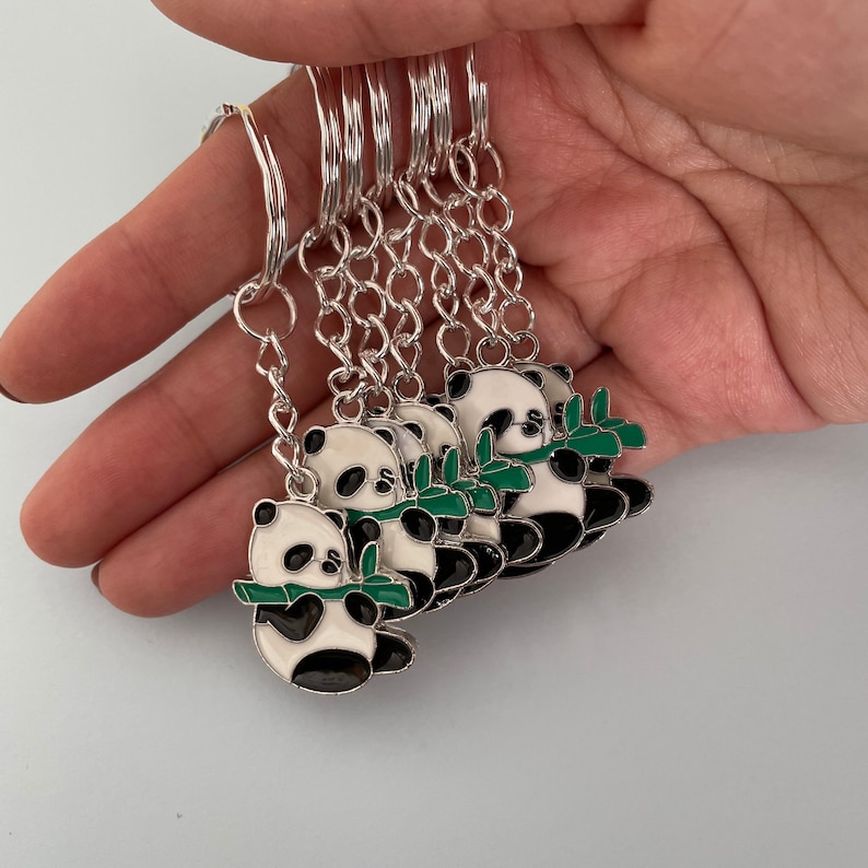 May include: A set of six silver keychains featuring panda bears holding green bamboo shoots. Each panda is outlined in black and has white fur with black eyes and ears.