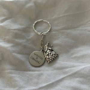 May include: Silver metal keychain featuring a round tag with the text "WINTER IS COMING" and a wolf head charm. The keychain has a ring for keys. The wolf head charm is detailed and textured.