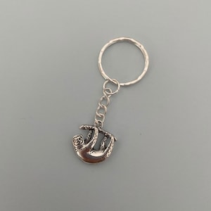 May include: Silver keyring featuring a sloth hanging from a branch. The keyring has a silver ring for attaching to keys.