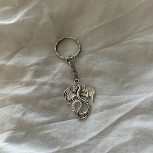 Peut inclure: Un porte-clés argenté en forme de dragon. Le dragon a des ailes détaillées et une queue enroulée. Le porte-clés comprend un anneau et une petite chaîne. Le fond est un tissu blanc texturé.