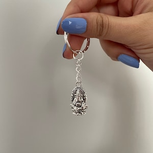 May include: Silver keychain featuring a detailed depiction of the Hindu deity Ganesha. The keychain has a silver ring and a silver chain.