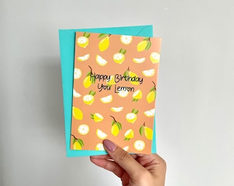 May include: A birthday card with a lemon pattern and the text "Happy Birthday You Lemon" is held in front of a teal envelope. The card features illustrations of whole and sliced lemons on a peach background.