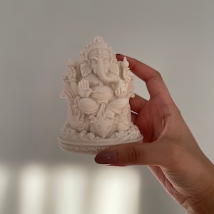 May include: White figurine of the Hindu deity Ganesha, sitting on a throne with a decorative base. The figurine is made of a smooth, white material and has intricate details.