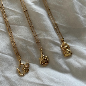 May include: Three gold-colored necklaces with different pendants. The pendants include the Om symbol with a small clear stone, a deity, and a figure with a crown. The necklaces are on a white fabric background.