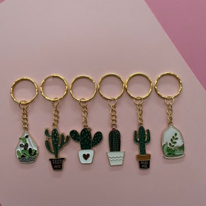 May include: Six gold keychains with different cactus designs. The keychains feature a cactus in a pot, a cactus with a heart, a cactus with the text "Feed Me", a cactus with the text "Hug Me", a cactus in a glass dome, and a cactus with a heart.