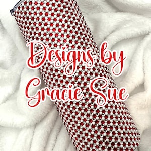 Może przedstawiać: Cylindryczny kubek pokryty czerwonymi i białymi kryształkami. Tekst "Designs by Gracie Sue" jest napisany czerwoną kursywą z przodu. Kubek ma czarną pokrywę i jest umieszczony na białym, puszystym tle.