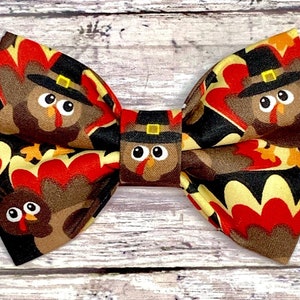 May include: A fabric bow tie featuring a repeating pattern of cartoon turkeys wearing pilgrim hats. The bow tie has a black background with red, brown, and yellow accents, perfect for Thanksgiving or fall celebrations.
