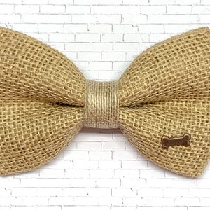 May include: A tan burlap bow tie with a bronze dog bone charm. The bow tie is made of woven burlap fabric and has a natural, rustic appearance. The bow tie is centered with a wrapped, tan-colored band.