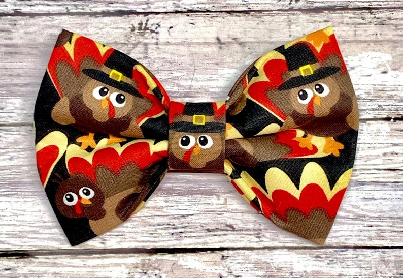 May include: A fabric bow tie featuring a repeating pattern of cartoon turkeys wearing pilgrim hats. The bow tie has a black background with red, brown, and yellow accents, perfect for Thanksgiving or fall celebrations.