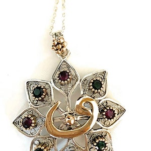 Op de afbeelding: Zilverkleurige hangerketting met een bloemmotief. De hanger heeft een goudkleurig Arabisch kalligrafiesymbool en is versierd met kleine, ronde, rode en groene edelstenen. De ketting heeft een fijne, zilveren ketting.