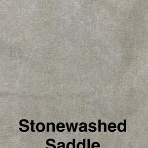 May include: Close-up of a textured, stonewashed fabric in a saddle color. The fabric has a subtle, mottled appearance, with variations in tone. The words "Stonewashed Saddle" are printed in black at the bottom of the image.