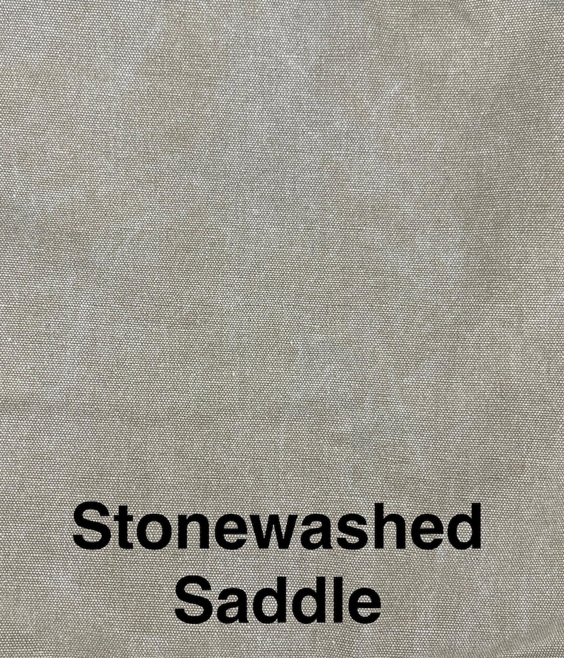 May include: Close-up of a textured, stonewashed fabric in a saddle color. The fabric has a subtle, mottled appearance, with variations in tone. The words "Stonewashed Saddle" are printed in black at the bottom of the image.