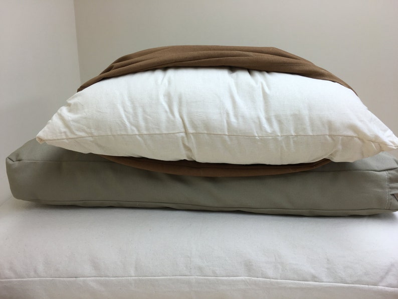 May include: A stack of three pillows in varying colors. The top pillow is covered with a brown fabric. The middle pillow is white, and the bottom pillow is a light gray. The pillows are stacked on top of each other.
