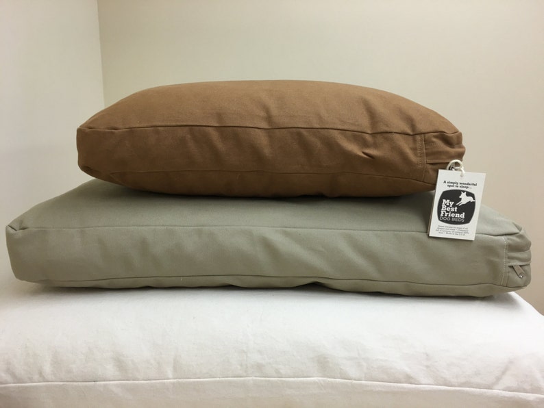 May include: Two dog beds stacked on top of a white pillow. The bottom bed is a light green color, and the top bed is brown. A tag that says "My Best Friend Dog Beds" is attached to the light green bed.