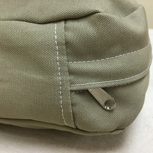 May include: Close-up of a khaki-colored canvas bag with a silver zipper and white stitching. The bag has a rectangular shape with a rounded top and a small handle. The canvas material has a textured appearance.
