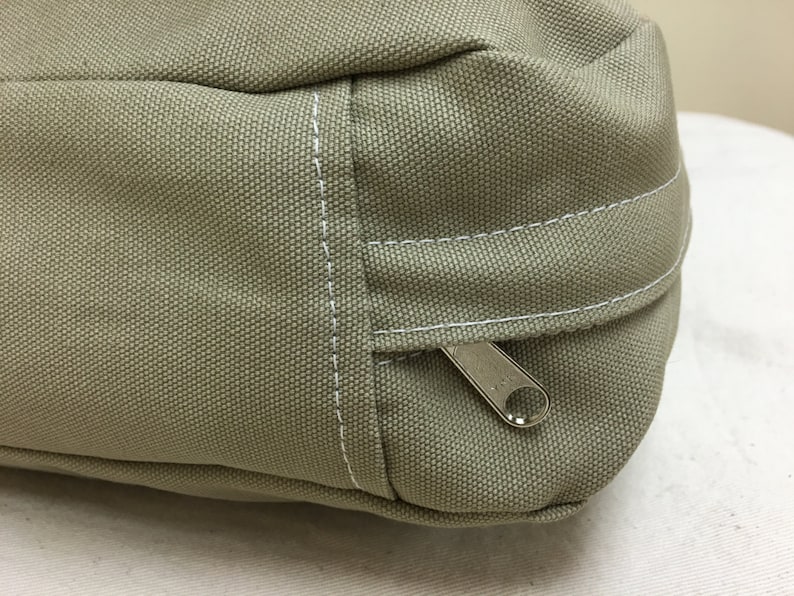 May include: Close-up of a khaki-colored canvas bag with a silver zipper and white stitching. The bag has a rectangular shape with a rounded top and a small handle. The canvas material has a textured appearance.