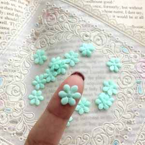 May include: A collection of small, light green flower-shaped embellishments. One flower is held on a finger, with others scattered on a white, lacy fabric. The background includes pages from a book.
