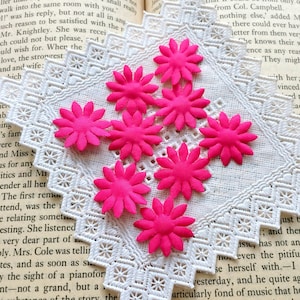 May include: Close-up of seven bright pink felt flower embellishments on a white doily. The flowers have a simple design with five petals and a small center.