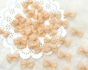 May include: A collection of small, pale peach-colored fabric bows. The bows are arranged on a white doily and scattered on a white fuzzy surface.