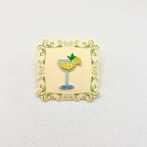 May include: An embroidered patch featuring a yellow cocktail in a light blue glass with a green garnish, centered on a square cream-colored card with a decorative green scrollwork border and scalloped edges, showcasing detailed stitching.