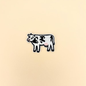 May include: Embroidered patch featuring a cartoon cow with black spots on a white body. The cow has a pink udder and is outlined in black. The patch is on a light beige background.