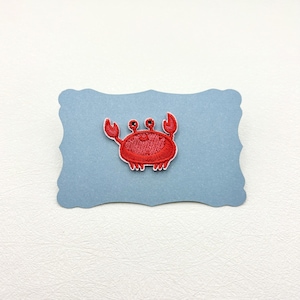 Peut inclure: Un écusson brodé en forme de crabe rouge vif avec deux yeux saillants, deux pinces levées et six petites pattes. Le crabe est bordé d'un contour blanc et est centré sur une carte rectangulaire bleu clair aux bords festonnés.